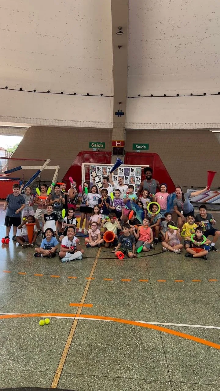 Grupo de crianças e educadores em quadra coberta com materiais esportivos e recreativos coloridos